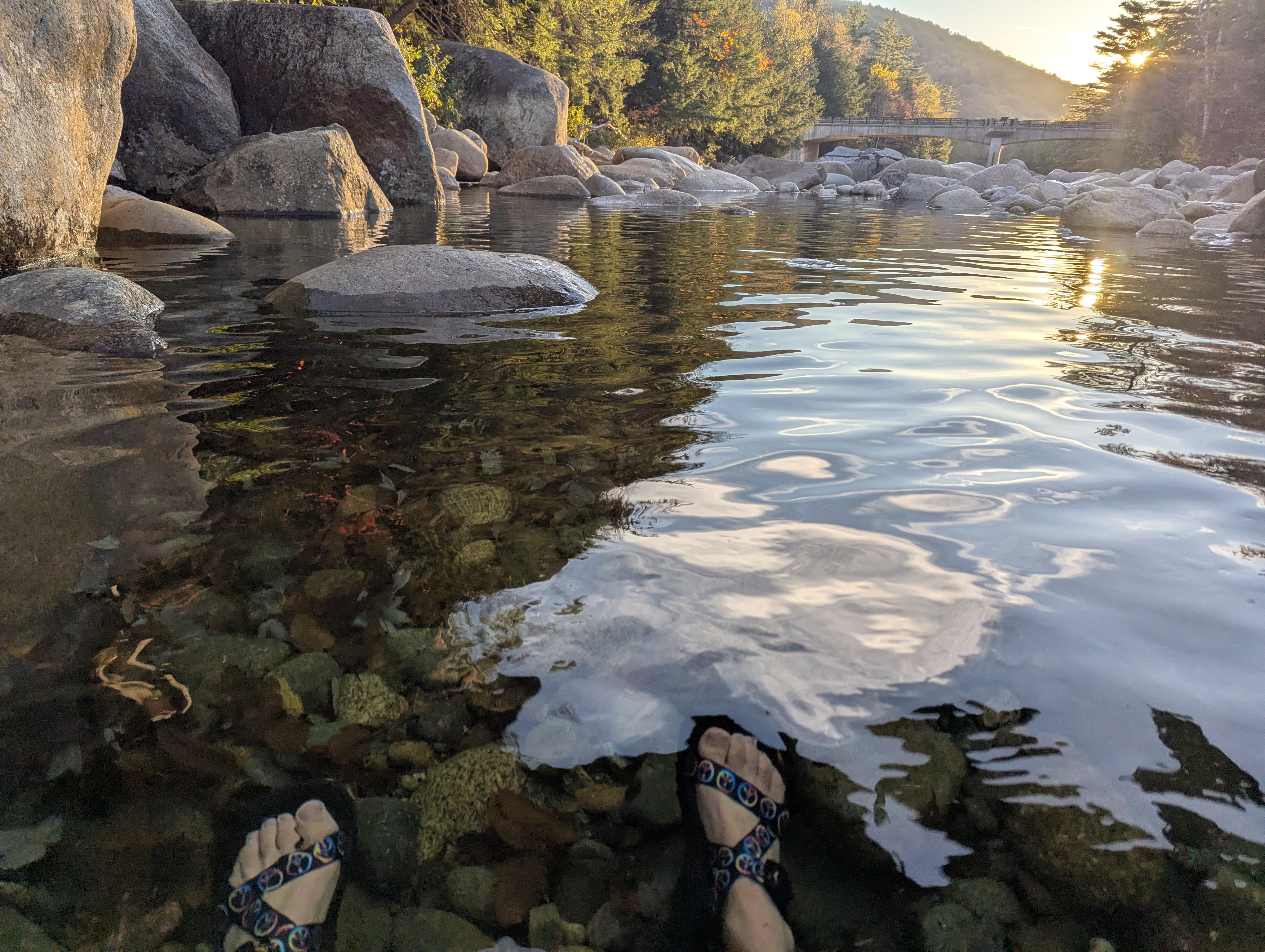 My feet in a clear river, with a concrete bridge in the distance and the setting sun behind it