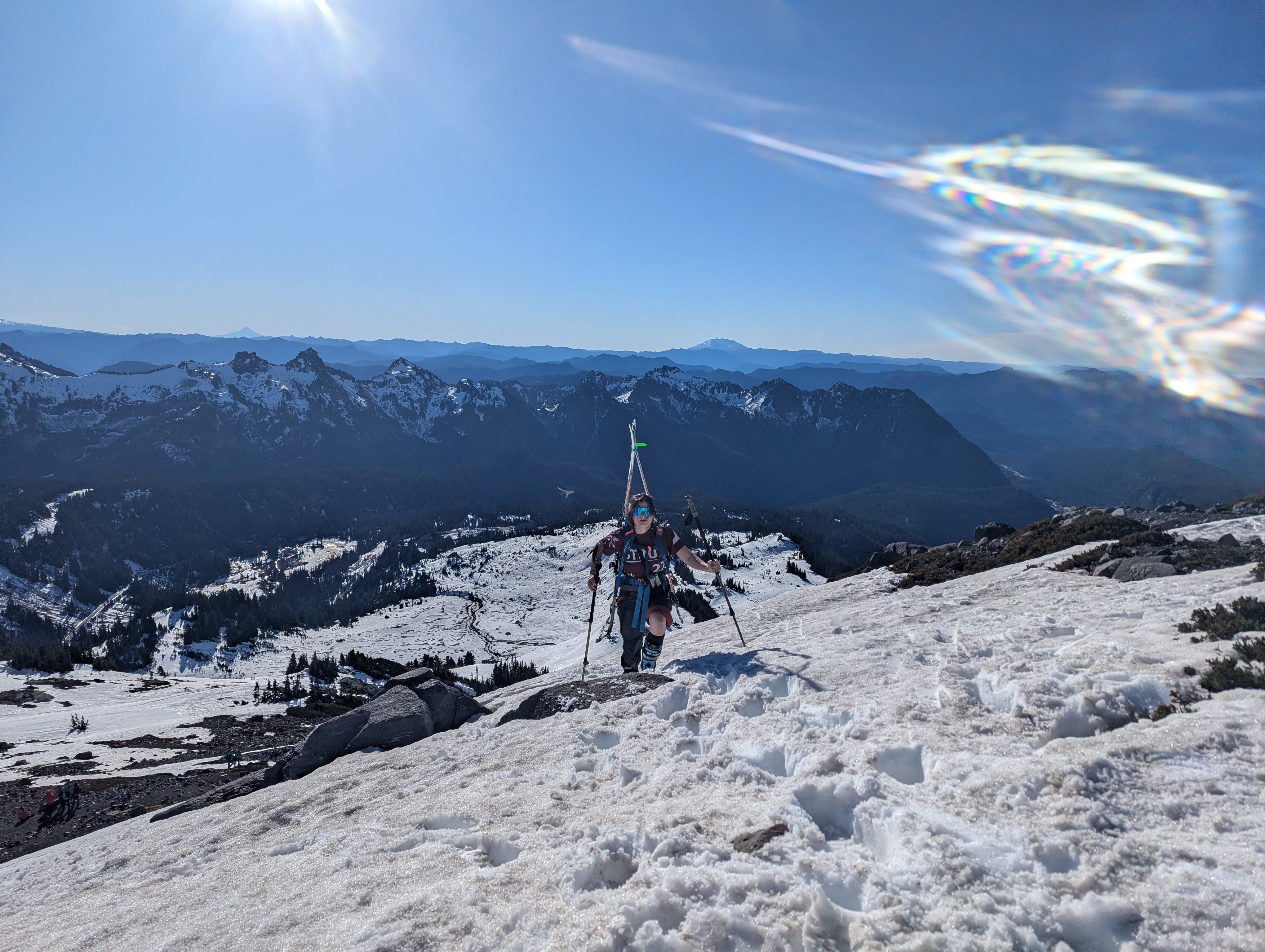 A skier carrying her skis ascending from behind a crest