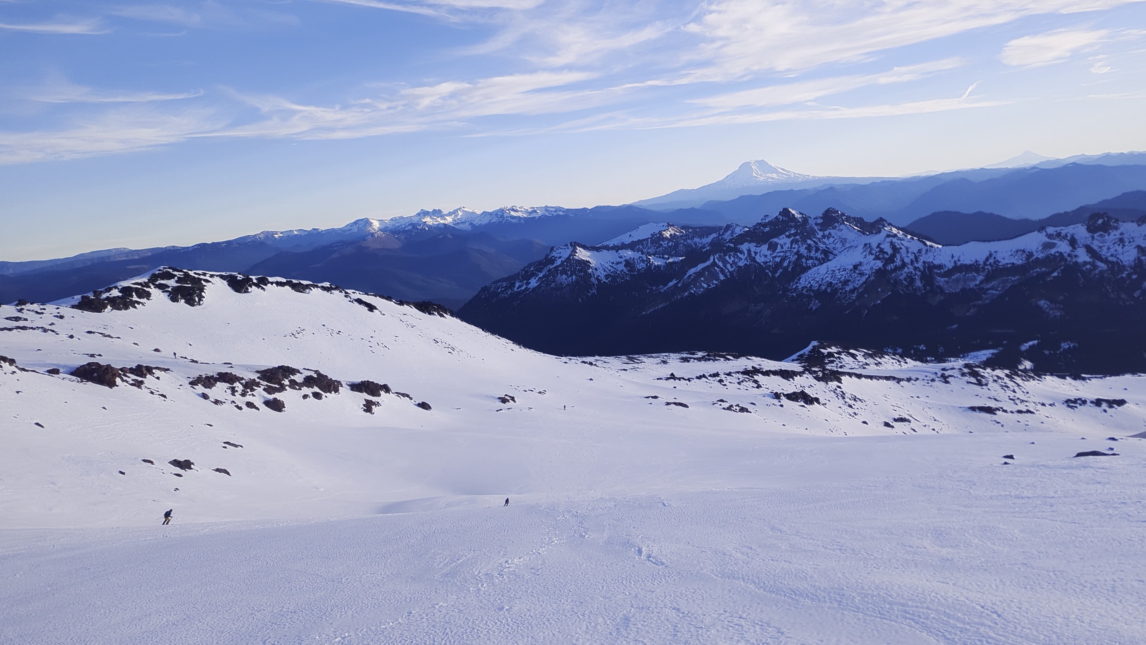 Small skiers on a wide open snow field, with more mountains in the background