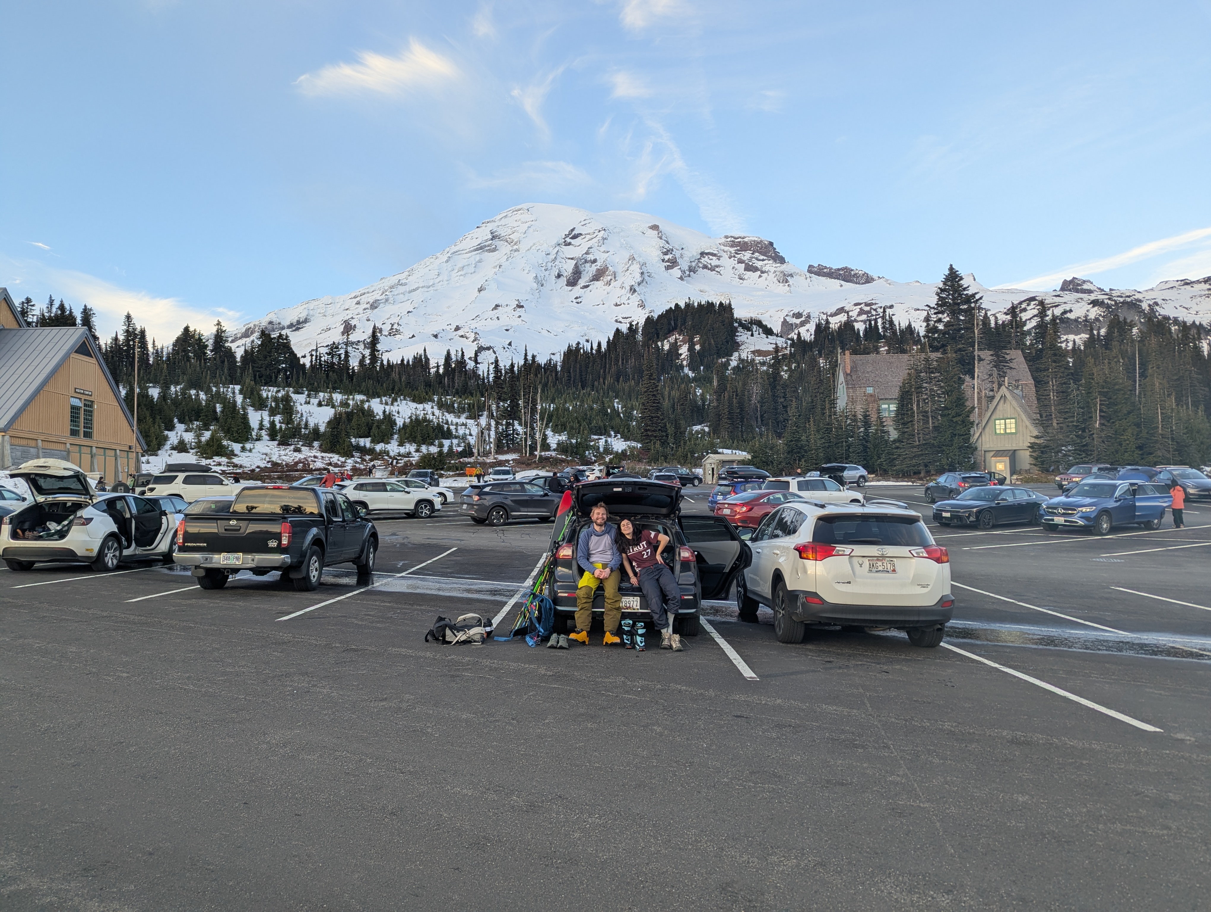 A couple sitting in the trunk of a car, ski gear strewn about, with a large mountain in the background