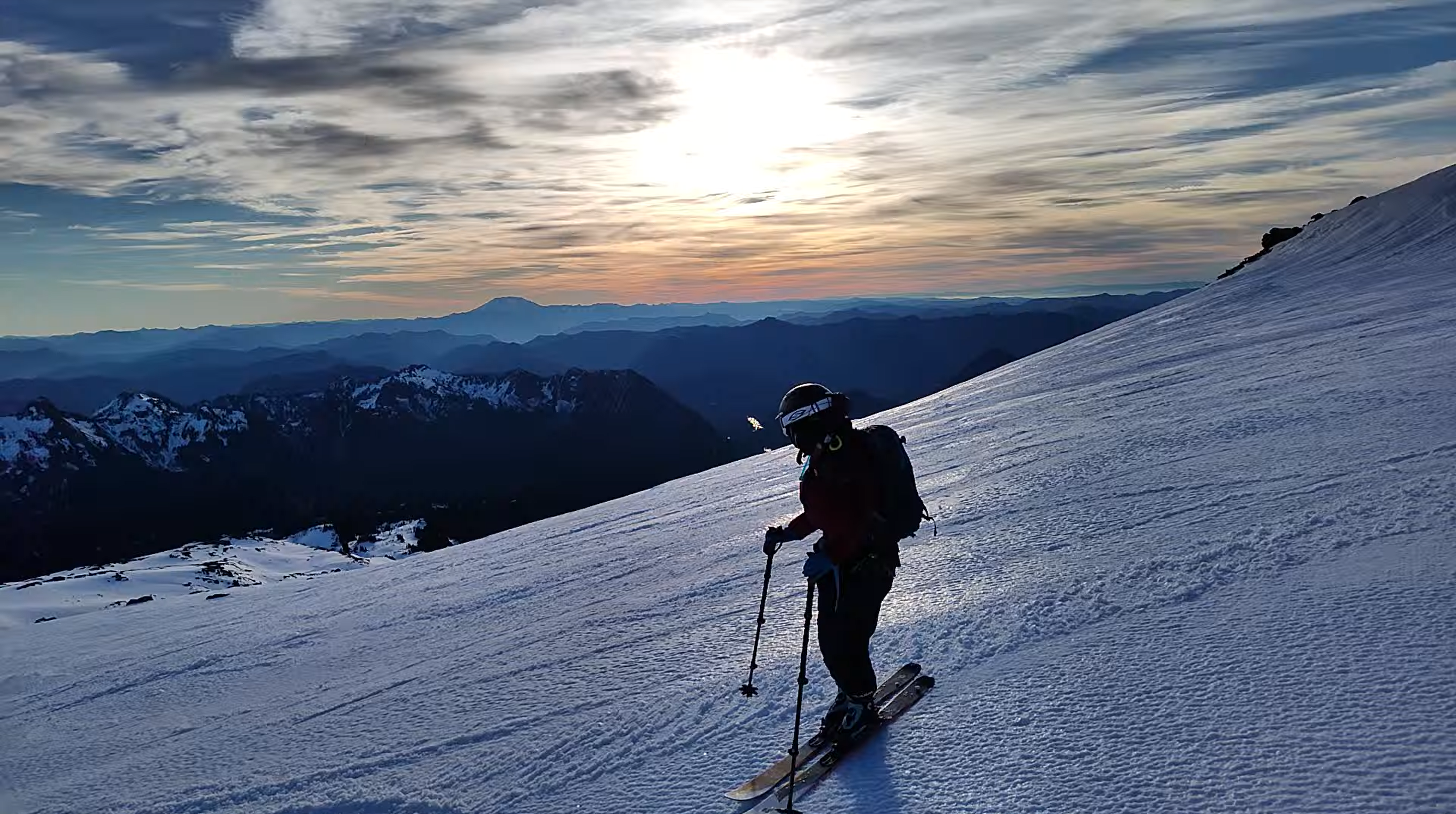 A skier pausing on a slope, with the setting sun a mountains behind